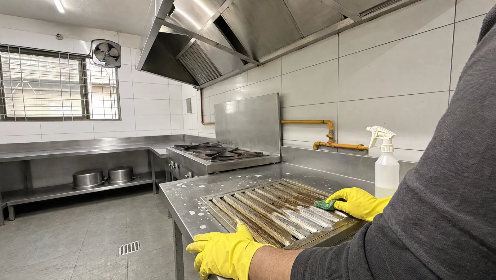 AI-generated illustration of gloved hands scrubbing grease from a kitchen hood filter in a stainless-steel staff kitchen.