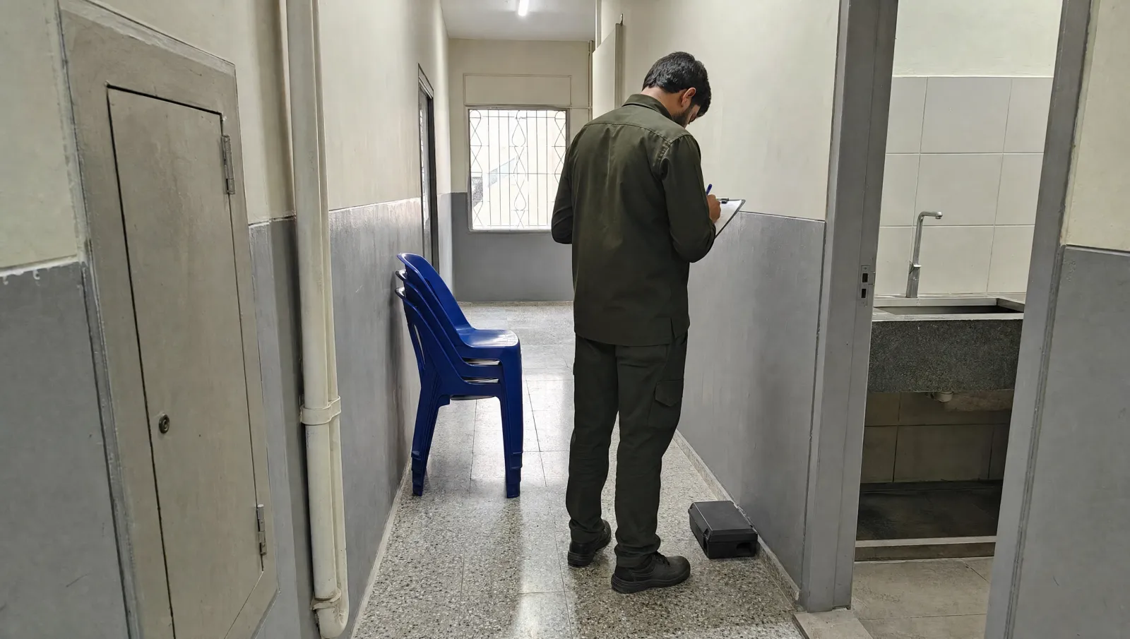 AI-generated illustration of a pest-control technician, seen from behind, recording a check beside a closed rodent bait station in an office service corridor.